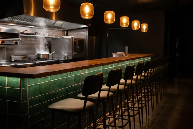 An empty restaurant counter late at night: green tiles, wooden top, empty stools, warm pendant lights and a little steam from a pot in the kitchen behind